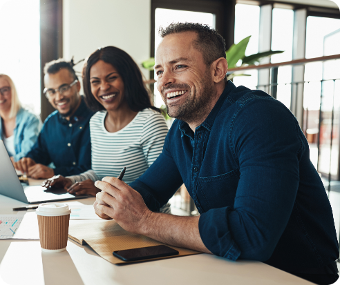 Four professionals around a table in a bright, modern office, with a man in a blue shirt smiling and engaged in discussion, alongside a woman in a striped shirt, creating a collaborative and inviting atmosphere.
