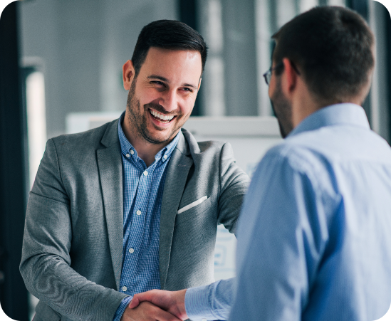 Two men in a professional setting engaging in a handshake, symbolizing collaboration. The first man, wearing a light gray blazer and blue checkered shirt, smiles warmly, while the second, in a light blue shirt and glasses, has a neutral expression, highlighting a positive business agreement.