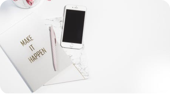 Minimalist workspace featuring a notebook with 'MAKE IT HAPPEN' embossed in gold, a sleek smartphone, a light pink polka dot pen, and a small container with pink flowers, conveying inspiration and productivity for mobile application development