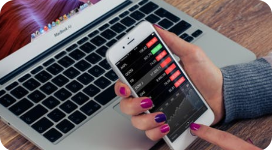 Close-up of a person's hand holding a smartphone displaying a stock market application with red and green price indicators, alongside a partially visible MacBook Air on a wooden surface, representing mobile application development in a finance-focused workspace.