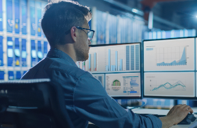 Person sitting at a desk in front of multiple monitors displaying colorful data visualizations and graphs, symbolizing data analysis, performance monitoring, and technology-driven decision-making in a high-tech office setting